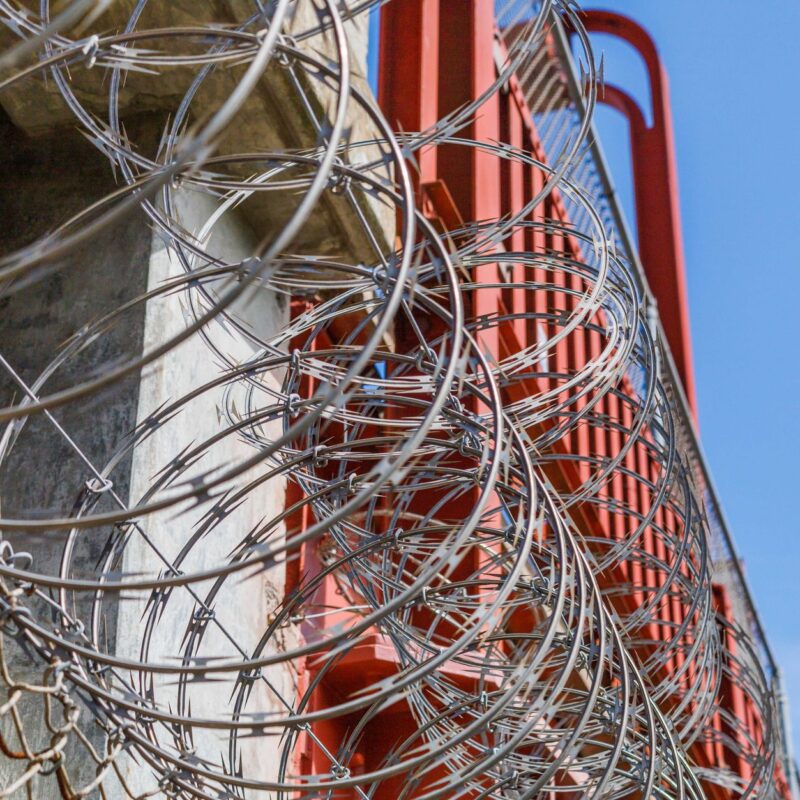 Im Vordergrund sind scharfe Rasiermesserdrahtspiralen zu sehen, die teilweise eine Betonstruktur und eine rote Metallkonstruktion verdecken, während im oberen rechten Bereich blauer Himmel sichtbar ist.