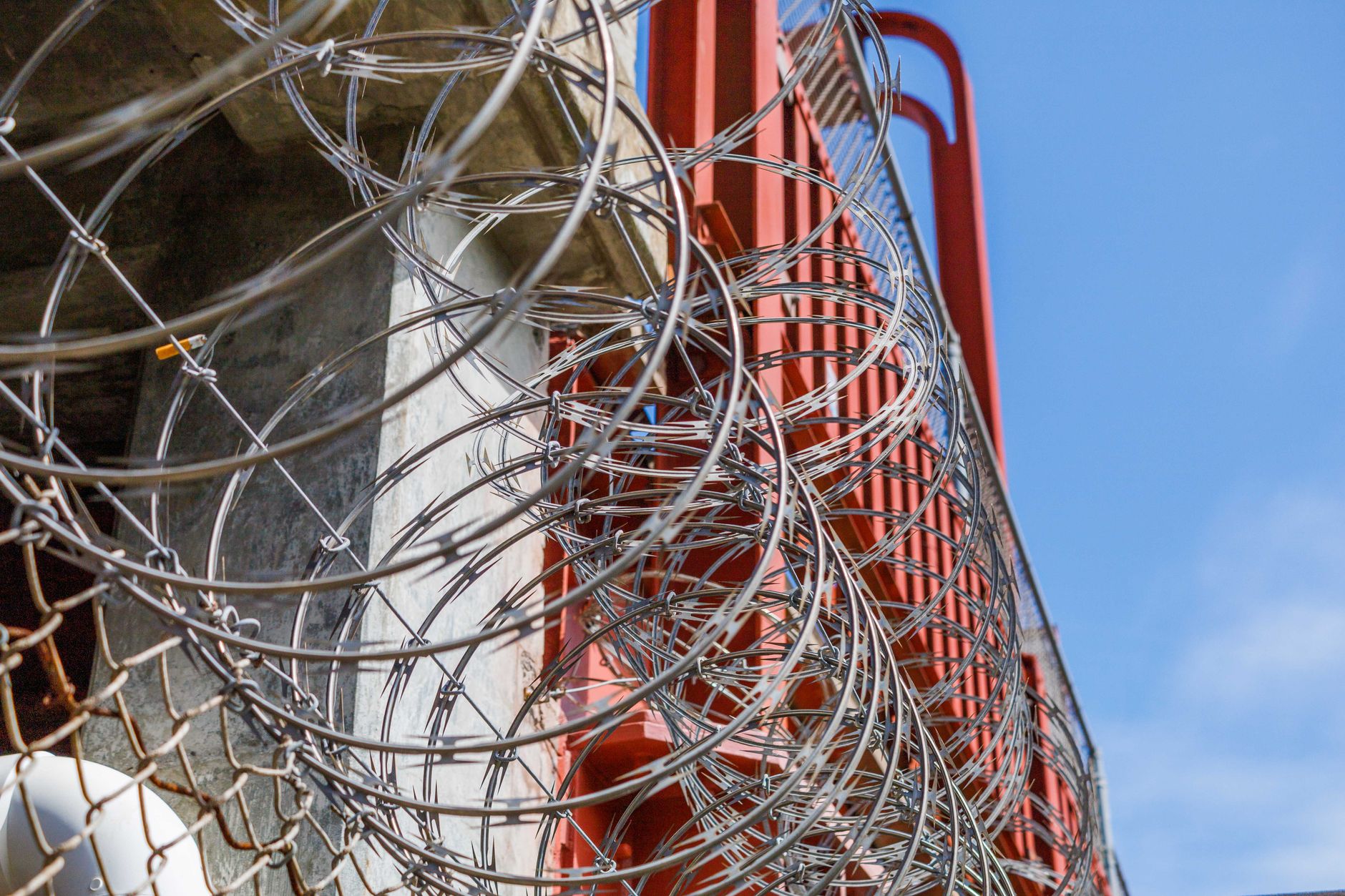 Im Vordergrund sind scharfe Rasiermesserdrahtspiralen zu sehen, die teilweise eine Betonstruktur und eine rote Metallkonstruktion verdecken, während im oberen rechten Bereich blauer Himmel sichtbar ist.