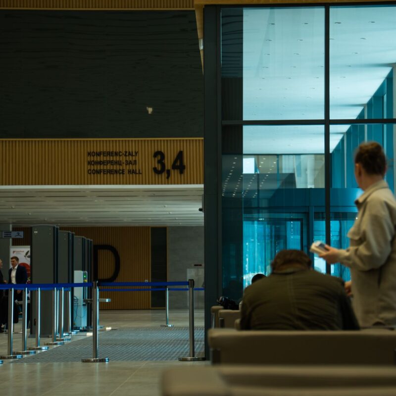 Ein moderner Innenraum mit einer Reihe von Sicherheitsschleusen und mehreren Personen. Im Hintergrund ist ein Schild mit der Aufschrift „CONFERENCE HALL 3,4“ zu sehen, während große Glasfenster auf der rechten Seite eine blaue Reflexion zeigen.
