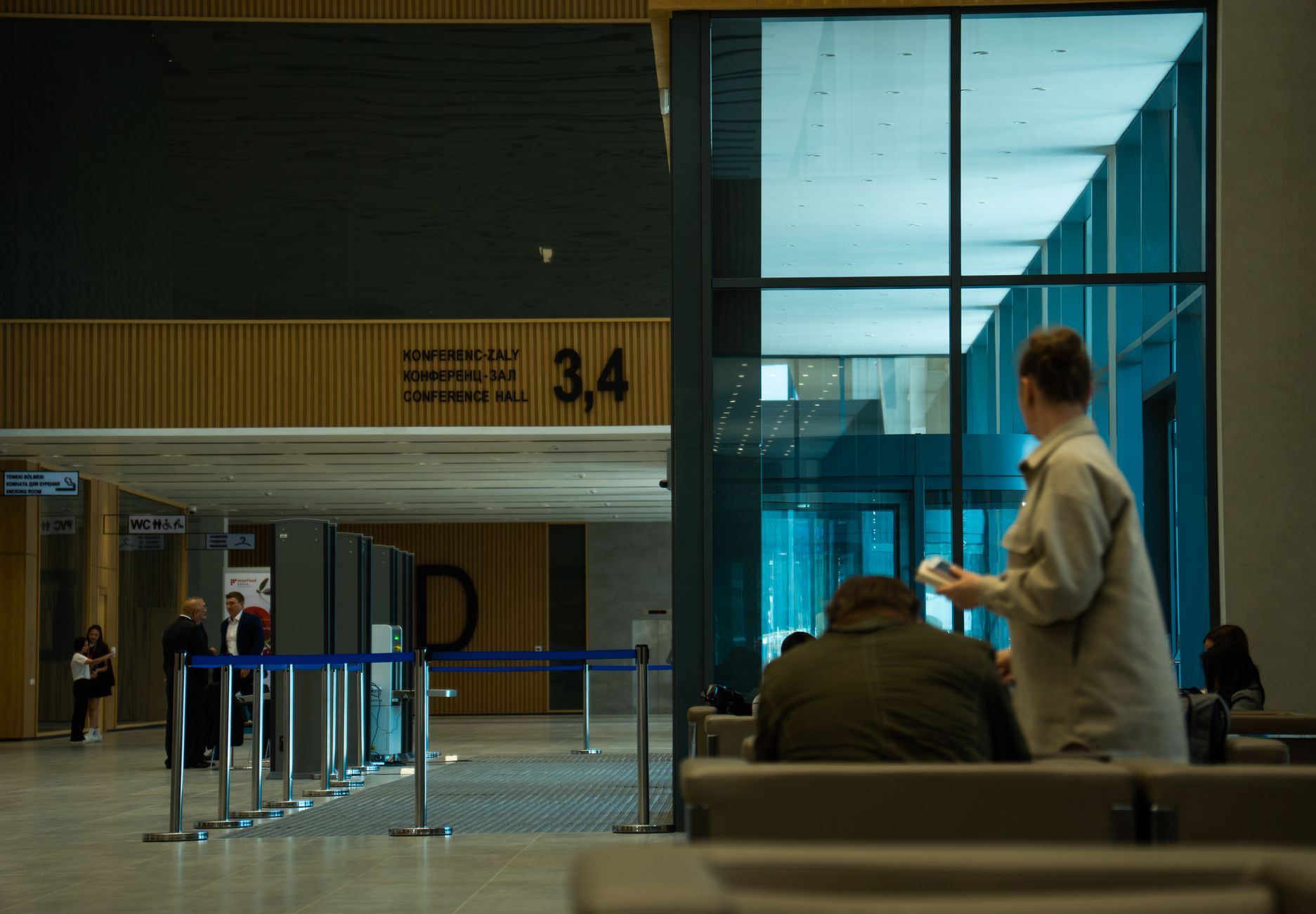 Ein moderner Innenraum mit einer Reihe von Sicherheitsschleusen und mehreren Personen. Im Hintergrund ist ein Schild mit der Aufschrift „CONFERENCE HALL 3,4“ zu sehen, während große Glasfenster auf der rechten Seite eine blaue Reflexion zeigen.