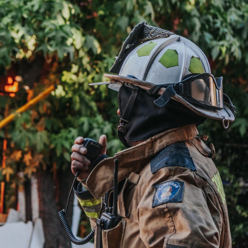 Ein Feuerwehrmann in brauner Uniform und Helm mit Schutzbrille hält ein Funkgerät an den Mund, während er vor einem Hintergrund aus grünen Blättern steht.