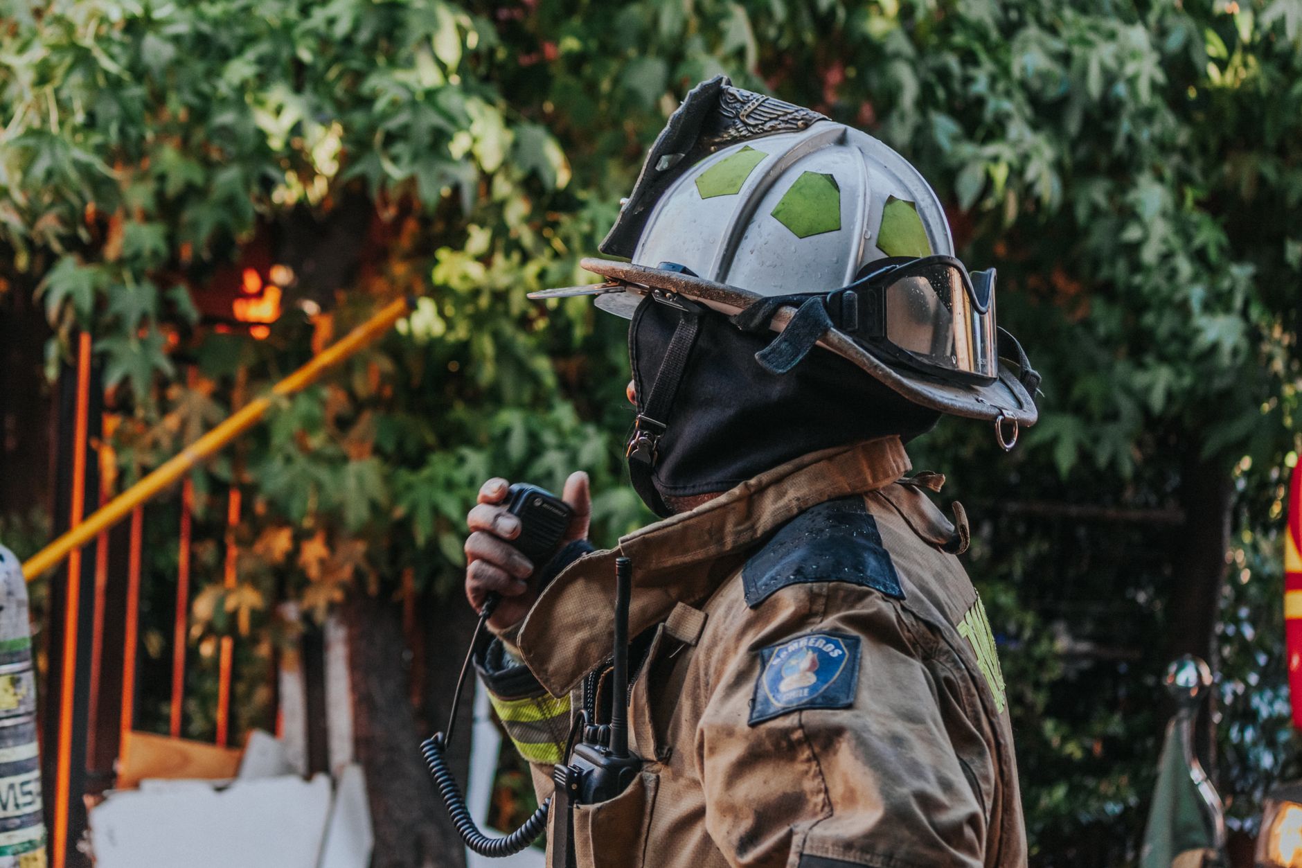 Ein Feuerwehrmann in brauner Uniform und Helm mit Schutzbrille hält ein Funkgerät an den Mund, während er vor einem Hintergrund aus grünen Blättern steht.