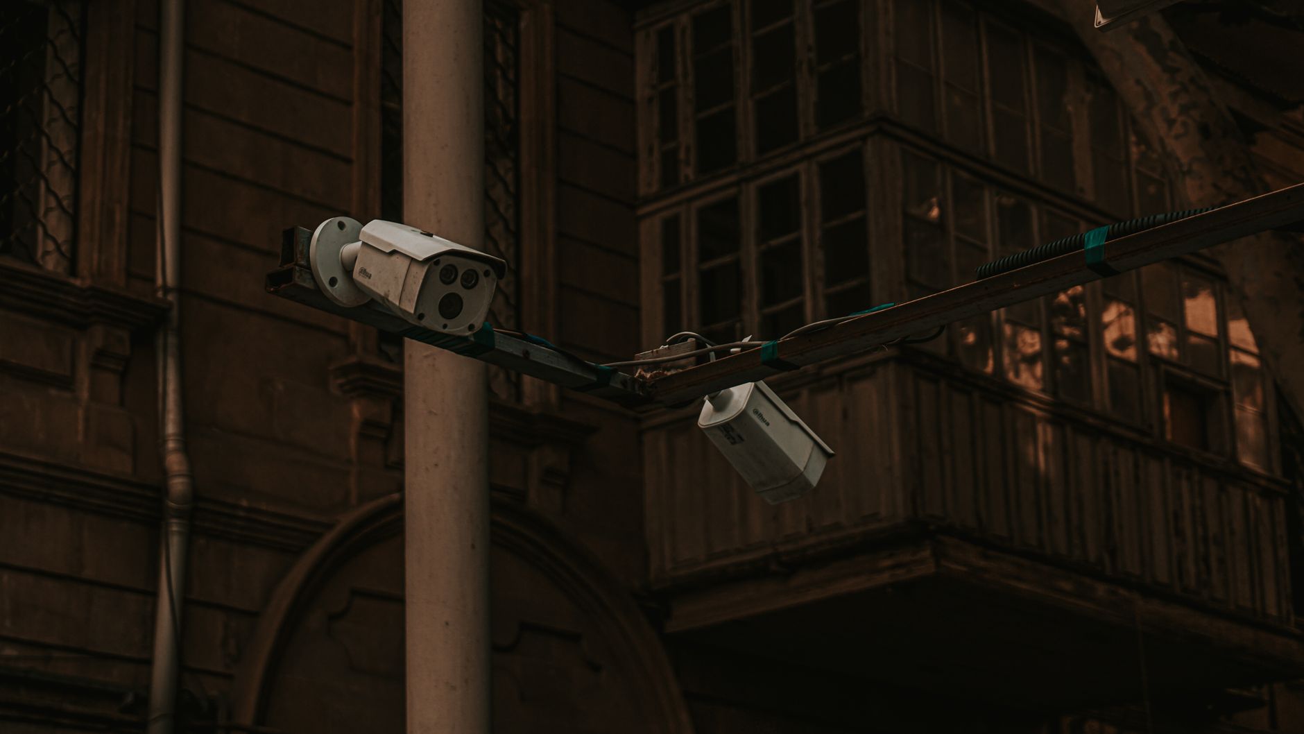 Zwei weiße Überwachungskameras sind an einem horizontalen Balken befestigt, der an einer hellen vertikalen Säule montiert ist. Im dunklen Hintergrund ist ein altes Gebäude mit Fenstern und einem Holzbalkon zu sehen.